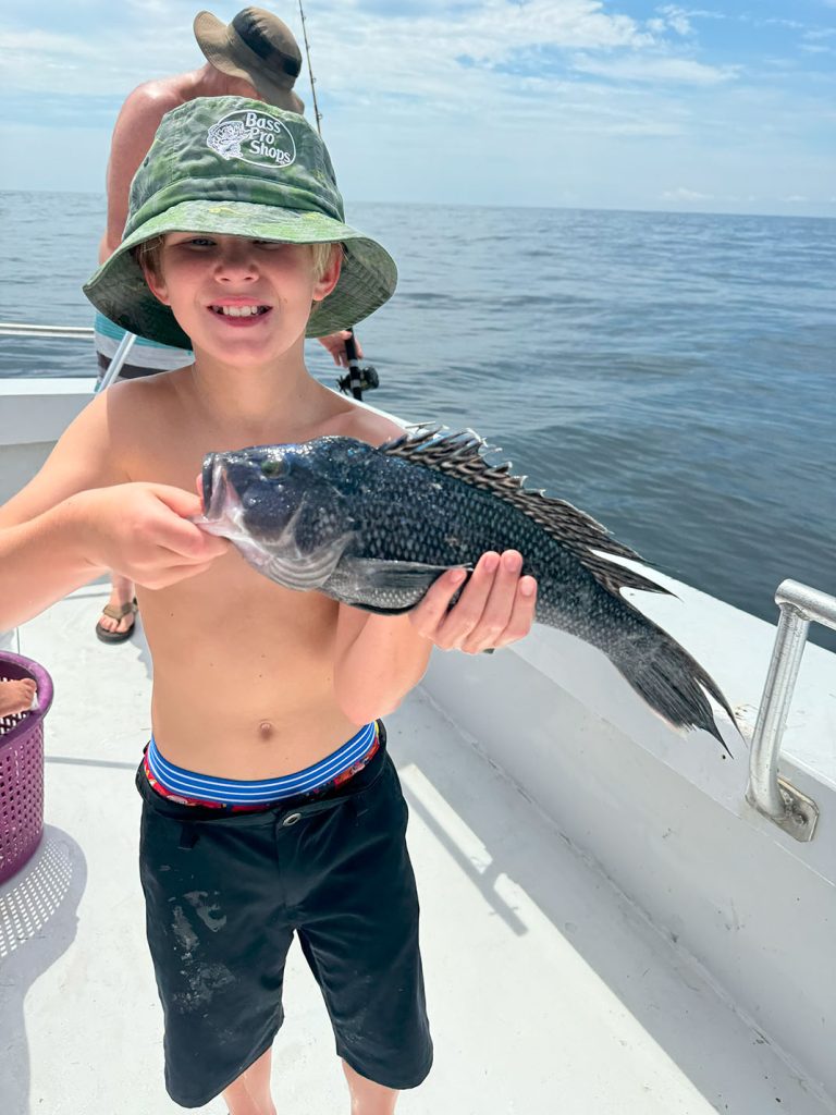 A young boy wearing a green hat and black shorts is smiling while holding a large fish on a fishing charter in Ocean City, MD. The ocean is in the background, and another person is fishing in the distance.