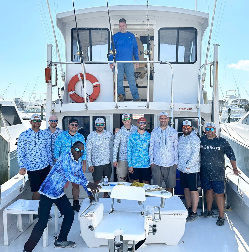 A group of ten people, mostly wearing sunglasses and patterned fishing shirts, stand on the deck of a fishing charter in Ocean City. One person is on the upper deck in blue. The sunny scene captures a lively marina with other boats visible in the background.