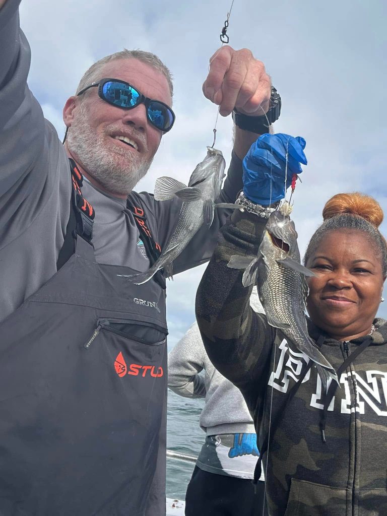 A man and a woman proudly display two fish they caught on a Fishing Charter in Ocean City, MD. The man wears a dark jacket and sunglasses, while the woman sports a camo hoodie and blue gloves. The sky is overcast, with the vast ocean providing an impressive backdrop.