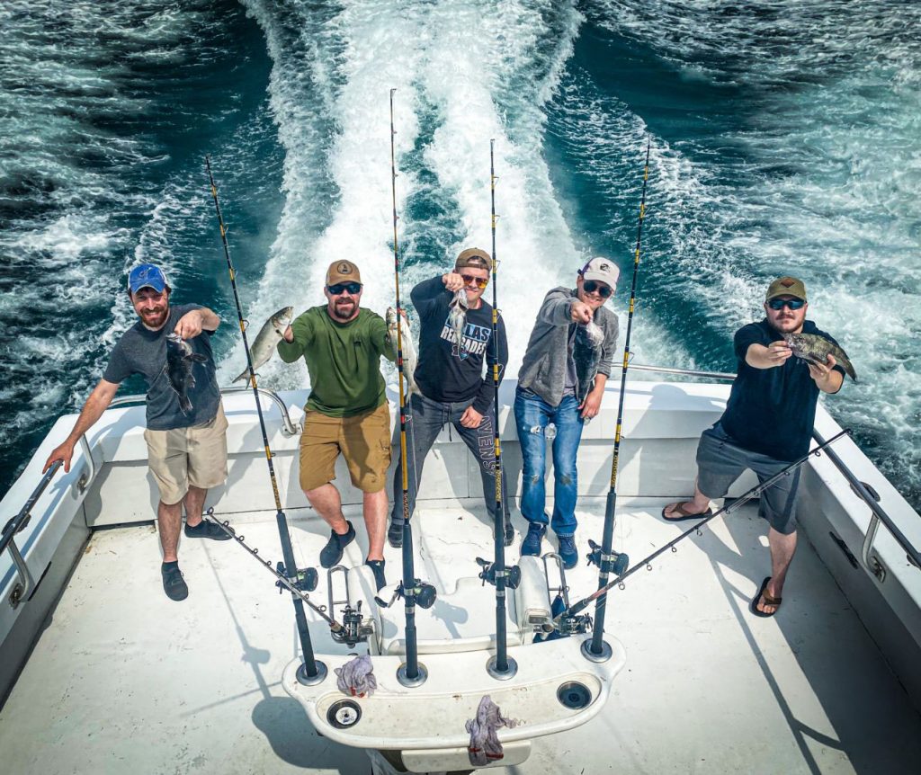 Five people on a Fishing Charter in Ocean City, MD, proudly hold up their catches, smiling at the camera. Fishing rods are neatly lined up in holders along the boat's railing as the wake creates a white foam trail on the blue water behind them.