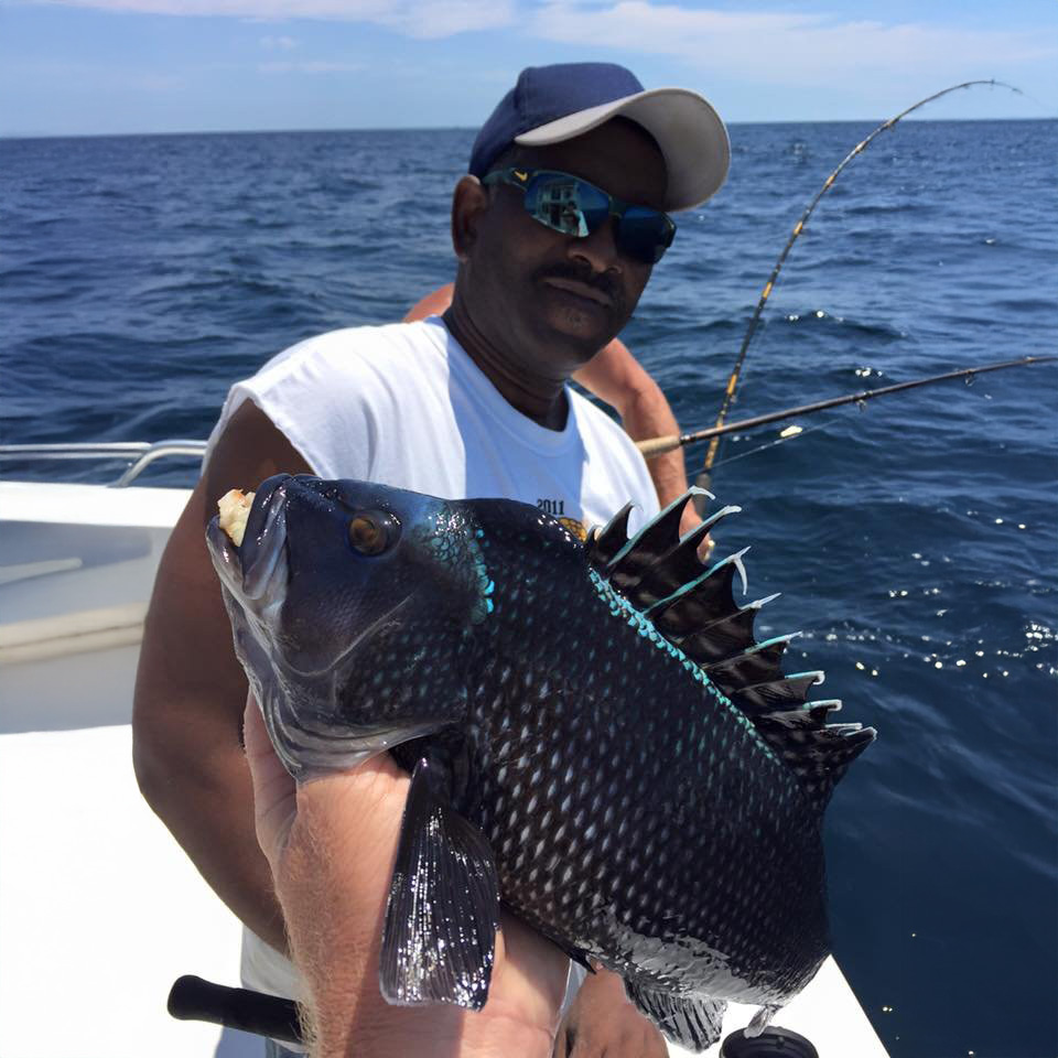 A person on a boat is proudly holding up a shiny, dark fish with sharp dorsal fins. Another person in sunglasses and a cap, perhaps part of the Fishing Charter Ocean City crew, stands in the background as the ocean off MD's coast glistens under a clear sky.