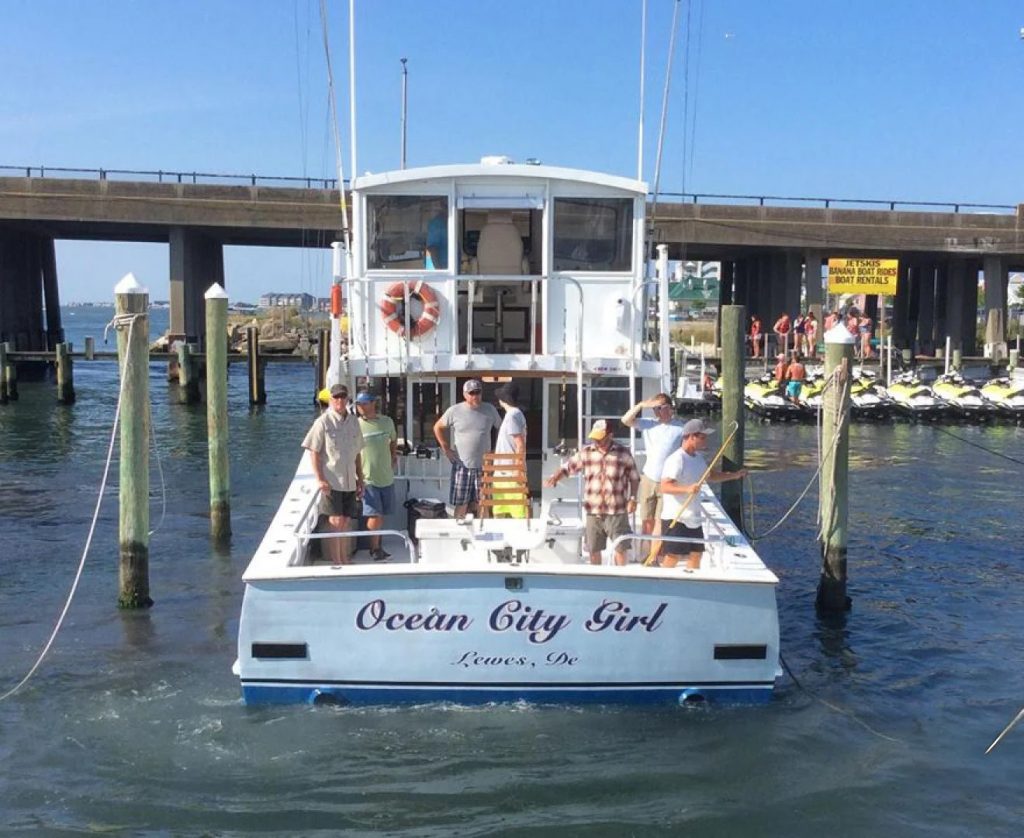 A boat named "Ocean City Girl" from Lewes, DE, reminiscent of the popular fishing charters in Ocean City, MD, is docked at a marina. Five people stand on the deck, some holding fishing rods. A bridge and several jet skis can be seen in the background.