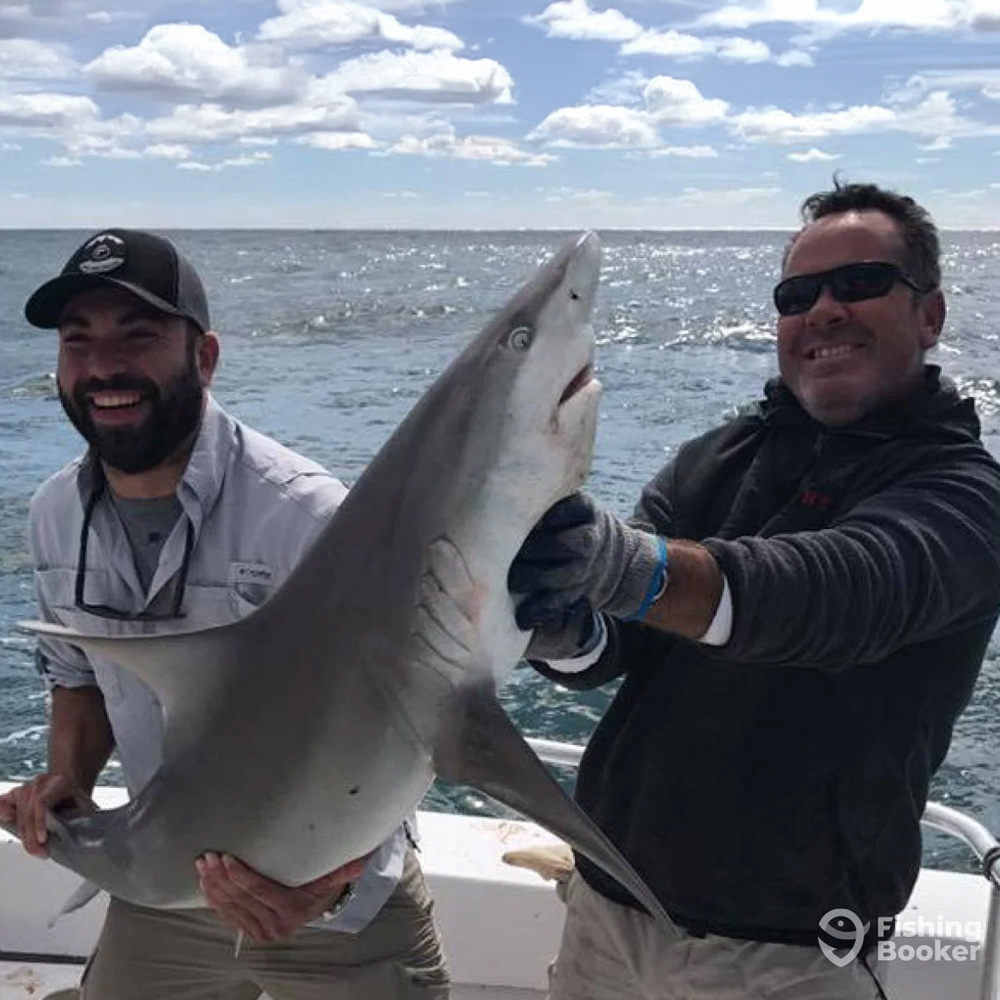 Two men smiling on a boat in Ocean City, MD, hold a small shark horizontally. The ocean stretches behind them under a partly cloudy sky. The man on the left wears a cap and the man on the right sports sunglasses. The "FishingBooker" logo is in the bottom right corner.