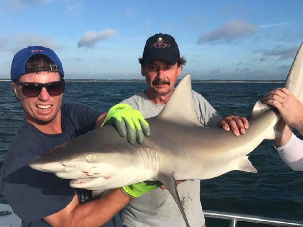 Two people on a fishing charter in Ocean City hold a small shark together on their boat, while a third person is partially visible. The ocean stretches behind them against a sky dotted with clouds, and both are dressed casually with one wearing gloves.
