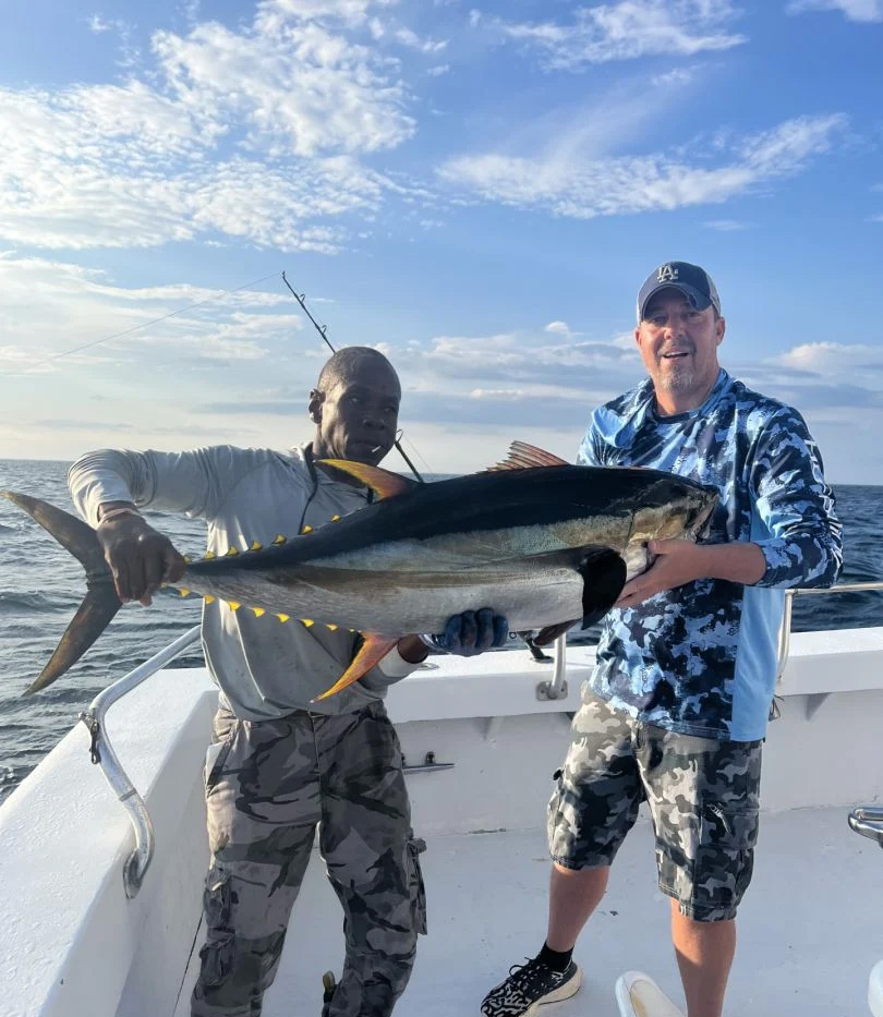 Two men on a boat with a large tuna, courtesy of an Ocean City fishing charter. The man on the left sports a light gray shirt and camo pants, while his MD companion on the right wears a blue camo hoodie and shorts. Behind them, the partly cloudy sky meets the vast ocean horizon.