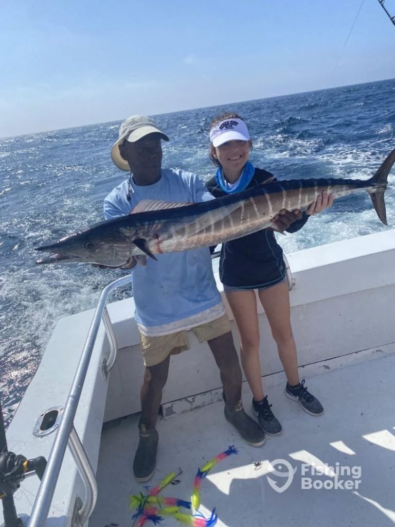Two people on a boat, part of an MD Fishing Charter Ocean City adventure, hold a large, striped fish against a backdrop of ocean waves. One wears a sun hat and shorts, the other a cap and blue neck gaiter. Colorful fishing lures are scattered across the boat's deck.