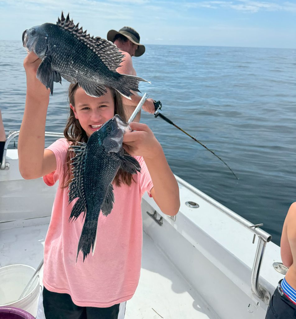 A smiling child in a pink shirt holds up two fish on a boat during a memorable fishing charter in Ocean City. An adult in a hat casts their line in the background. The ocean is calm under the clear Maryland sky.