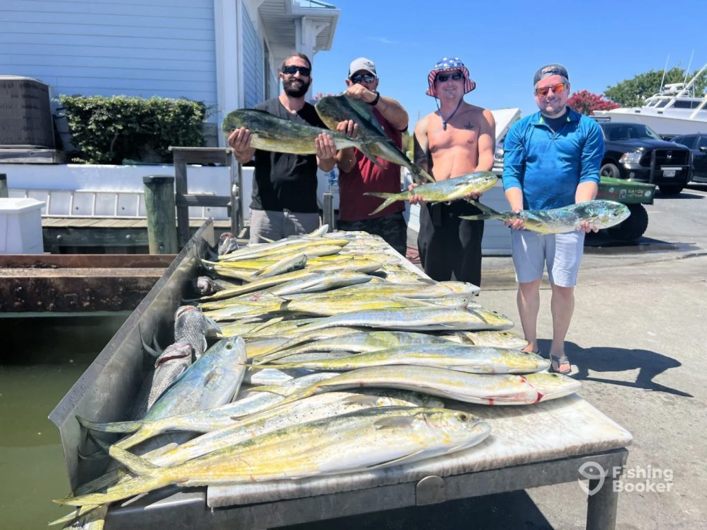 Four men proudly display their mahi-mahi catch from a Fishing Charter in Ocean City on a dockside table surrounded by freshly caught fish. Behind them, the white building and shimmering water set the scene. They're all smiles, dressed in casual summer attire with a star-spangled hat and sunglasses.