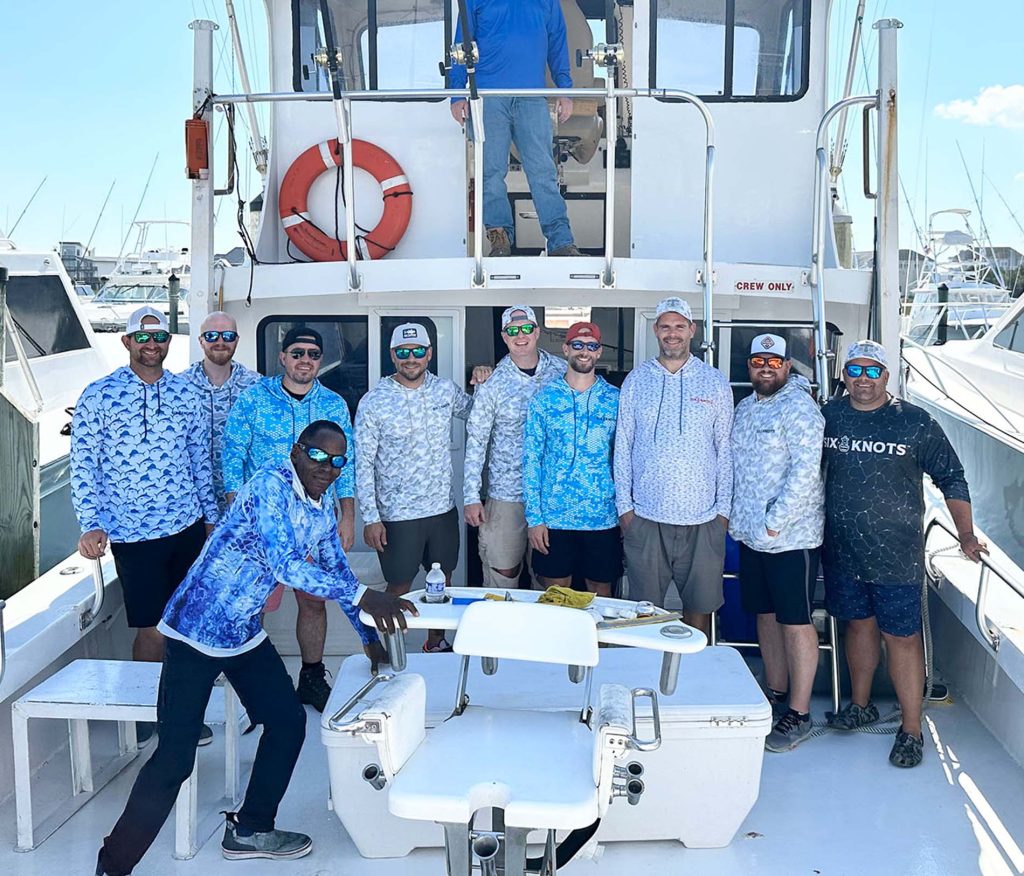 A group of ten men in fishing attire poses on a boat with gear visible, ready for their Ocean City fishing charter adventure. Docked at the marina, with other boats in the background, they smile widely, embodying the spirit of MD's vibrant fishing community.