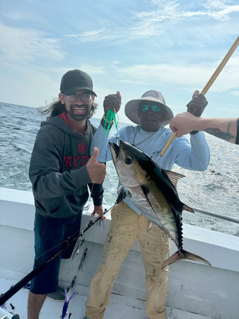 Two men on a Fishing Charter in Ocean City proudly display a large tuna they caught. One man, wearing a cap and glasses, gives a thumbs-up. The other, in a sunhat and reflective sunglasses, holds the fishing pole against the backdrop of the sparkling MD ocean under a partly cloudy sky.