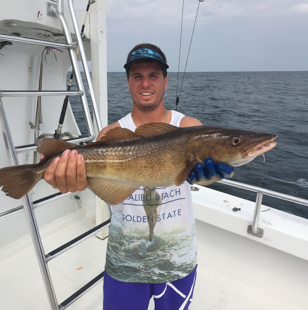A person on a boat proudly holds a large fish with both hands, celebrating a successful day with a Fishing Charter in Ocean City, MD. They're wearing a visor, sleeveless white shirt, and blue gloves. The ocean shimmers in the background, along with part of the boat’s railing and ladder.