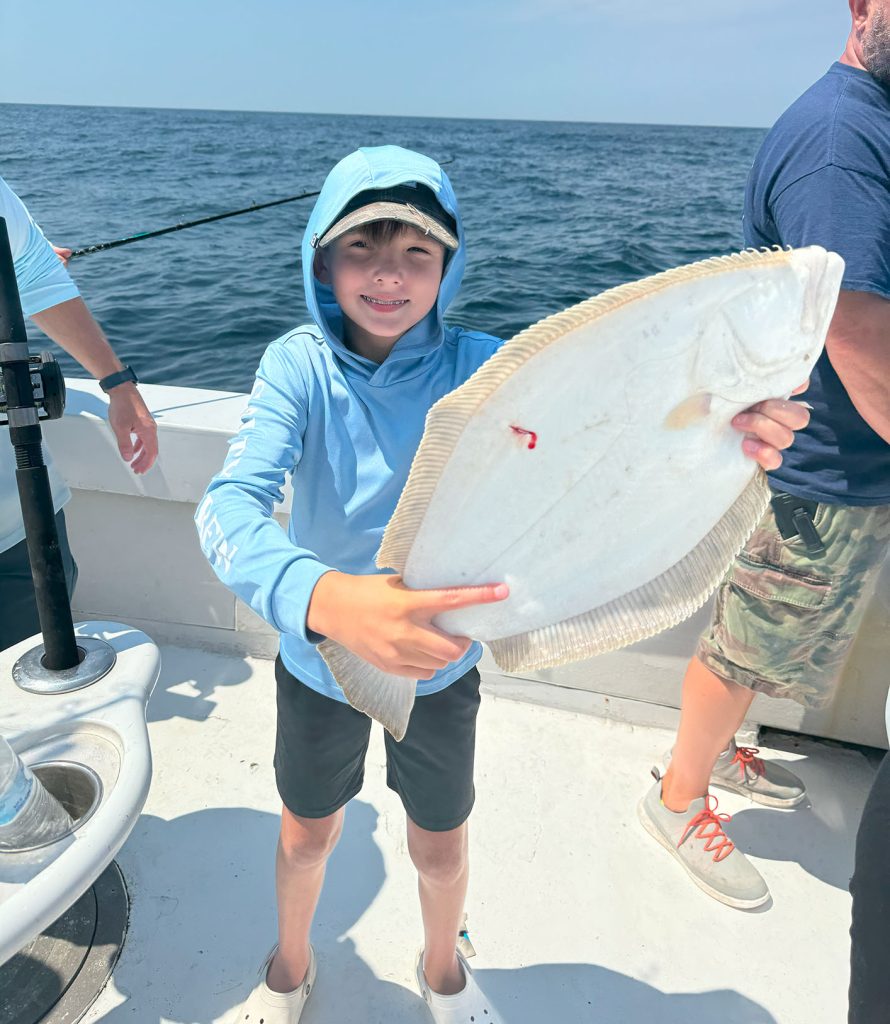 A child in a blue hoodie proudly holds a large flat fish aboard the Fishing Charter in Ocean City, MD. The sea stretches out in the background, and a few people are nearby, with one sporting camo shorts. It's a sunny day filled with adventure.