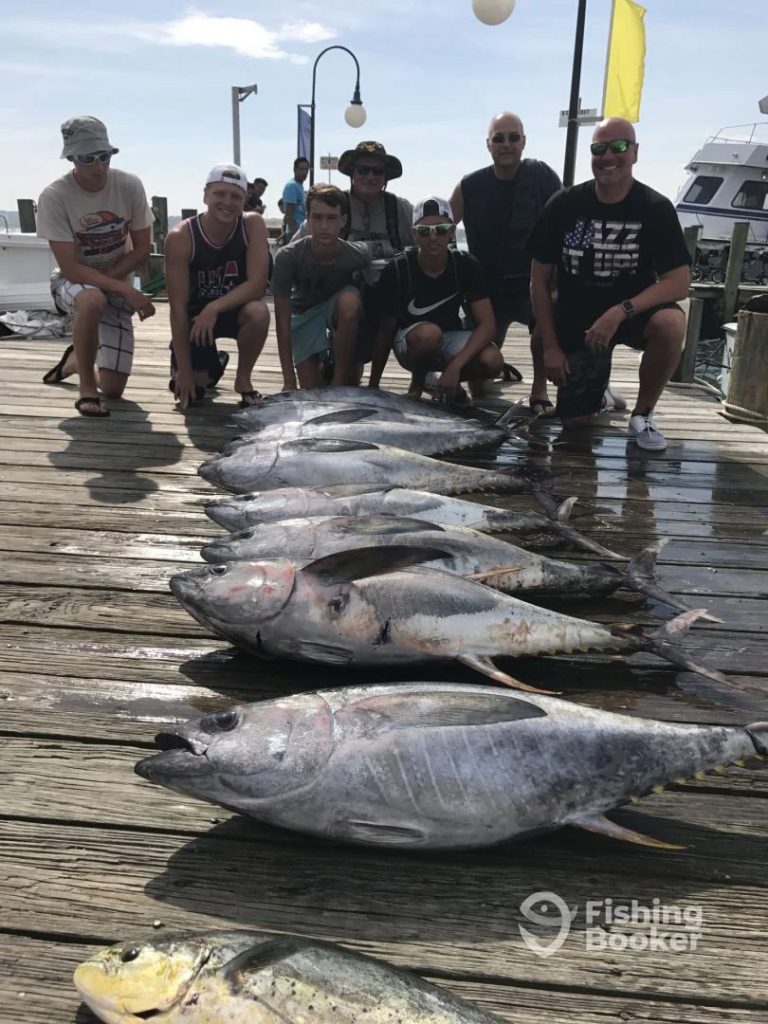 A group of eight people poses on a wooden dock in Ocean City, MD, with several large tuna laid out before them. The sun is shining, and a boat equipped for a fishing charter awaits in the background. A "Fishing Booker" logo graces the bottom right corner.