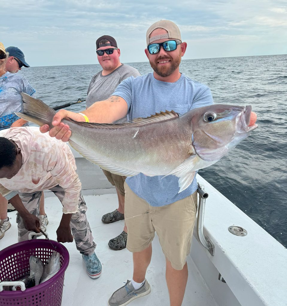 A smiling person on a boat holds a large fish proudly, showcasing the success of their Fishing Charter in Ocean City, MD. They sport sunglasses and a cap. In the background, others are busy, with one sorting fish into a purple basket against the backdrop of the ocean and an overcast sky.