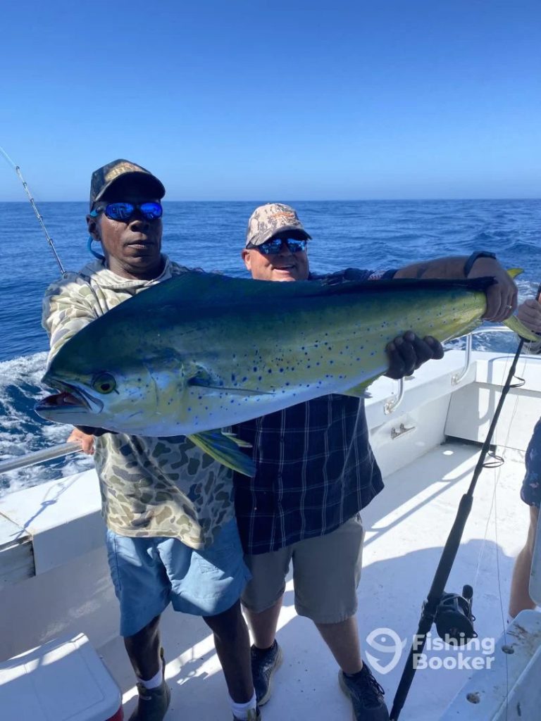 Two people on a fishing charter in Ocean City, MD, proudly hold a large mahi-mahi aboard their boat. They're both wearing sunglasses and hats, with the ocean stretching endlessly behind them under a clear blue sky.