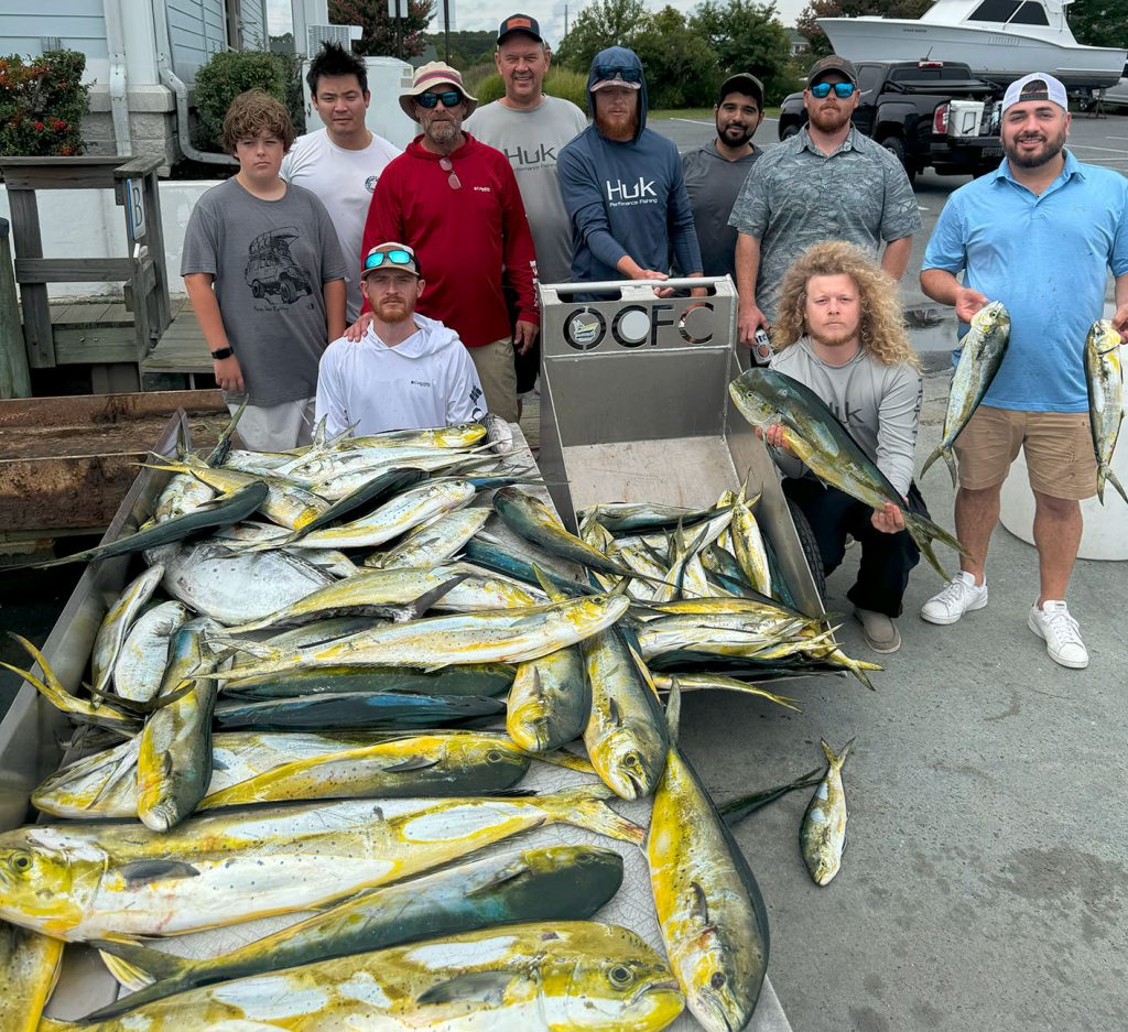 A group of nine people proudly pose with a large haul of fish, including mahi-mahi, on a cart after an exhilarating day with a Fishing Charter in Ocean City, MD. Two members hold fish triumphantly. They're outdoors near a dock, with a small boat and lush trees completing the scenic backdrop.