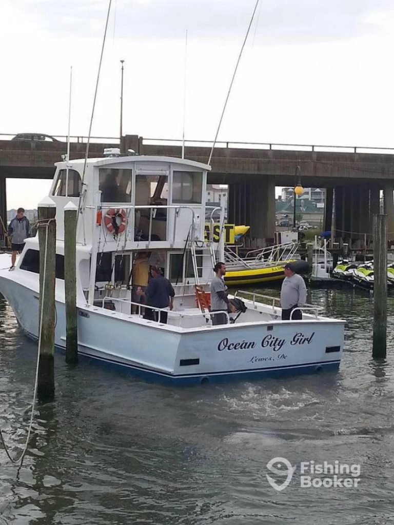 A white boat named "Ocean City Girl" is docked near a bridge in a marina, offering a perfect spot for an MD fishing charter. Four people are aboard, with jet skis and a yellow-and-black boat visible in the background under the cloudy sky.