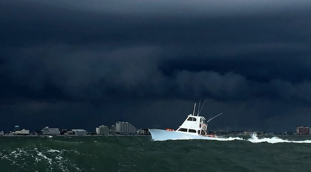 A boat, part of a Fishing Charter in Ocean City, navigates through rough, teal waters under a dark, stormy sky. The horizon reveals MD's distant city skyline with tall buildings silhouetted against the ominous clouds. The boat creates white waves as it moves forward.