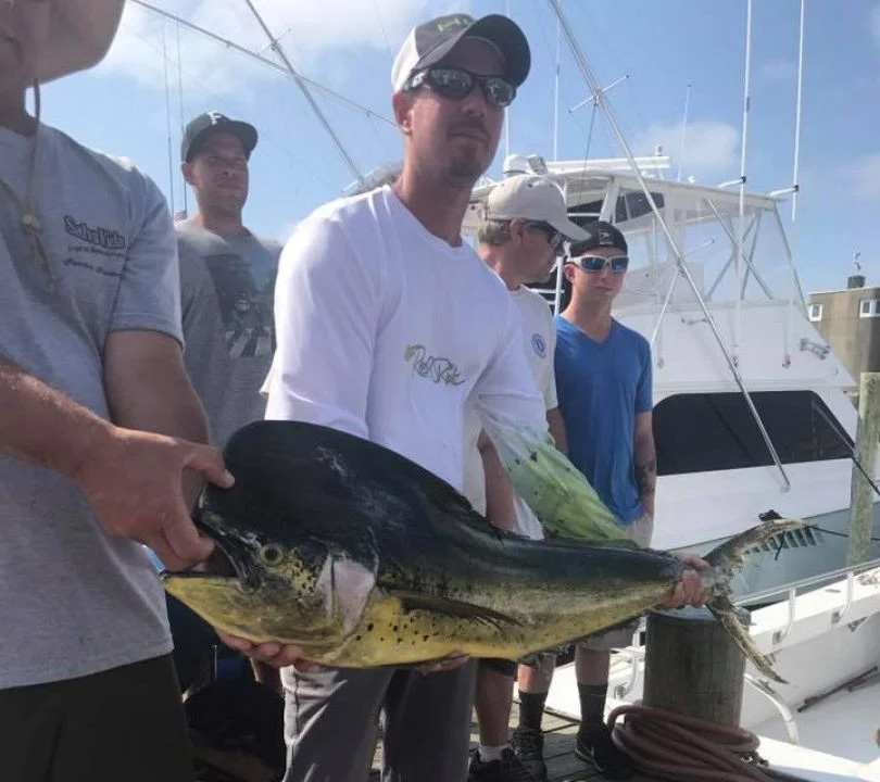 A group of men stands on a dock, two proudly holding a large mahi-mahi caught during their Fishing Charter Ocean City adventure. They’re near a boat, dressed in casual attire under the clear sky.