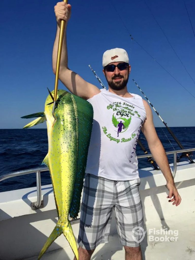 A man on a boat proudly displays a large fish caught with a spear. Sporting sunglasses, a white sleeveless shirt, plaid shorts, and a cap, he's the picture of success in Ocean City's renowned Fishing Charter. The ocean and several fishing rods stand as testament to his day of triumph.