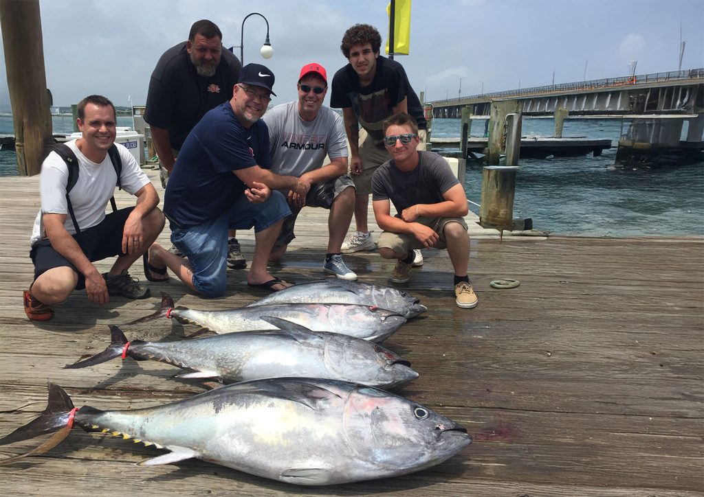 Six men, part of a Fishing Charter in Ocean City, are posing on a wooden dock in MD with four large, caught fish laid out in front of them. A bridge spans the waterway in the background under a partly cloudy sky.