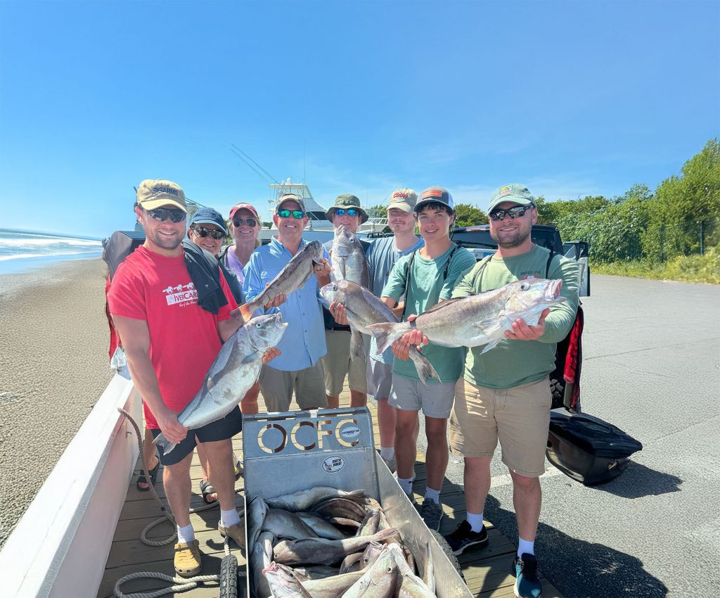 A group of people stand on a fishing charter boat near Ocean City, MD, holding large fish and smiling at the camera. The boat is close to a beach on a sunny day, with a container labeled "OCFE" filled with more fish in the foreground.